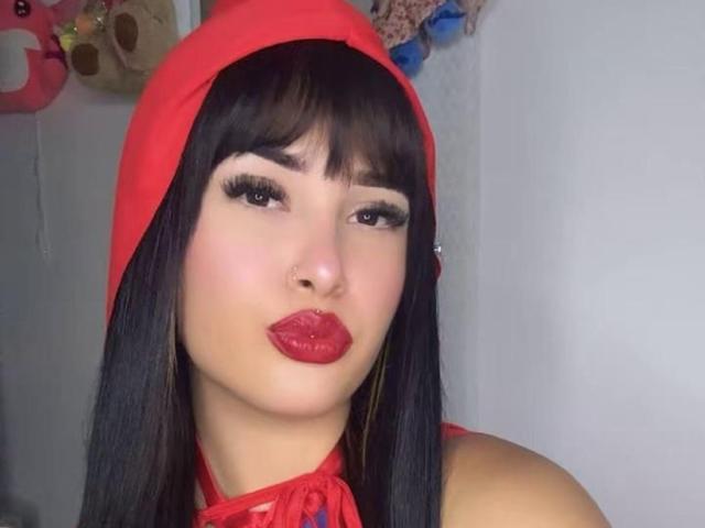 LunaAndKlaus has dark straight hair, a red hood, red lipstick, and long eyelashes, with a pouty expression, wearing a red halter top, and golden earrings.