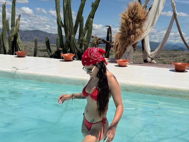 PrincessMiley stands in a pool, wearing a red bikini and a red bandana with white patterns. She has long dark hair and wears sunglasses. Her right arm, with tattoos, is raised. Cacti and potted plants are in the background, with a hammock draped on the right. The clear water contrasts with her tan skin. Mountains are visible under a blue sky with white clouds. She's in a desert-like setting.