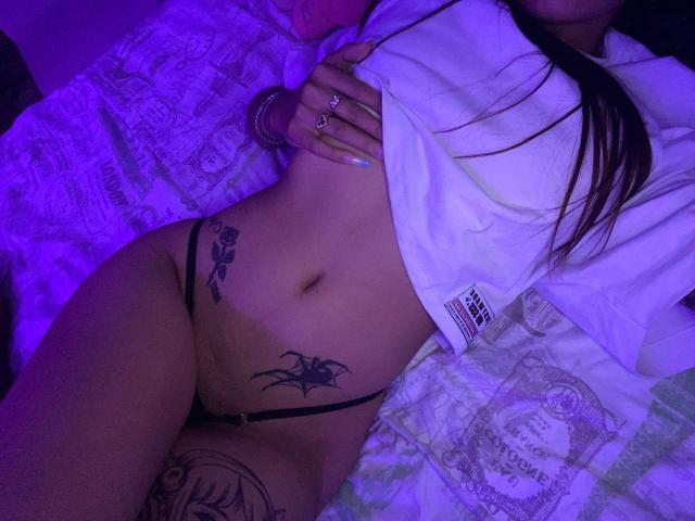 PrincessMiley lies on a bed with purple lighting. She has a white shirt pulled up, revealing a tattooed lower abdomen with a black thong. Her right hand, adorned with rings, rests on her chest. She has tattoos on her right hip and lower stomach. Her long brown hair is partially visible. The bed has a patterned quilt. The image focuses on her midsection and tattoos.