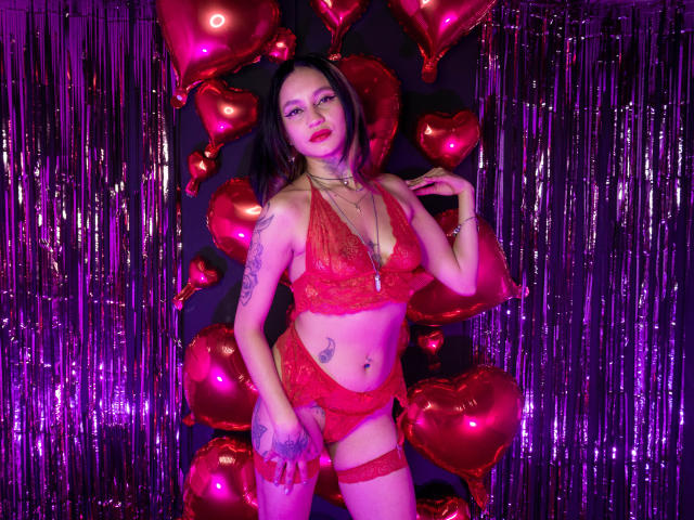LiiaSmith with red lace lingerie, tattoos, and garter, standing in front of shiny purple and red heart balloons, posing with hand on hip, looking at camera, smiling.