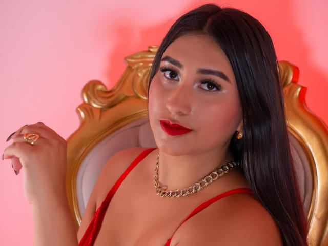 Catalina Rivero, com longos cabelos pretos, batom vermelho e vestido vermelho, sentada em uma cadeira dourada, usando um colar de corrente dourada, segurando um anel, sorrindo sutilmente. Seios visíveis..