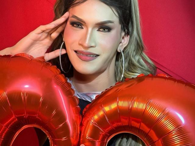 AgathaRodriguez, with light brown hair and green eyes, holds shiny red balloon letters "LOVE" against a red background. She wears large silver hoop earrings, has light pink lipstick, and a subtle smile. Her right hand touches her cheek. The balloons have a metallic texture, reflecting light. Her makeup is precise, with dark eyeliner and well-defined eyebrows. The composition focuses on her upper body and face. The image uses bright, contrasting colors and emphasizes the shiny, reflective quality of the balloons.