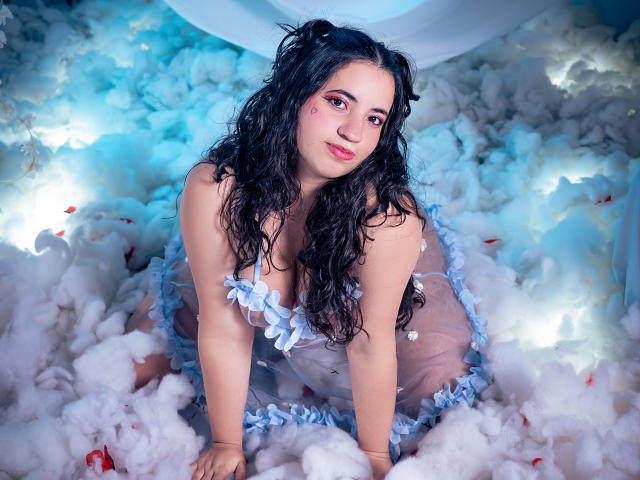 NicolSky, avec ses cheveux noirs bouclés, sa robe à fleurs bleues et son rouge à lèvres rose, est agenouillée dans une mousse blanche et moelleuse, montrant sa poitrine, son visage mignon et son petit tatouage en forme de cœur sur sa joue droite..