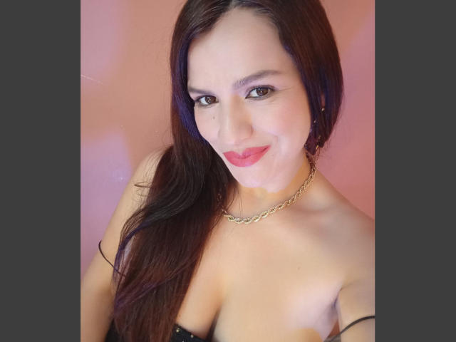 CameronViviane has red lipstick, brown eyes, and long brown hair, smiling at the camera, wearing a black strap top and gold chain necklace, with visible small tits.