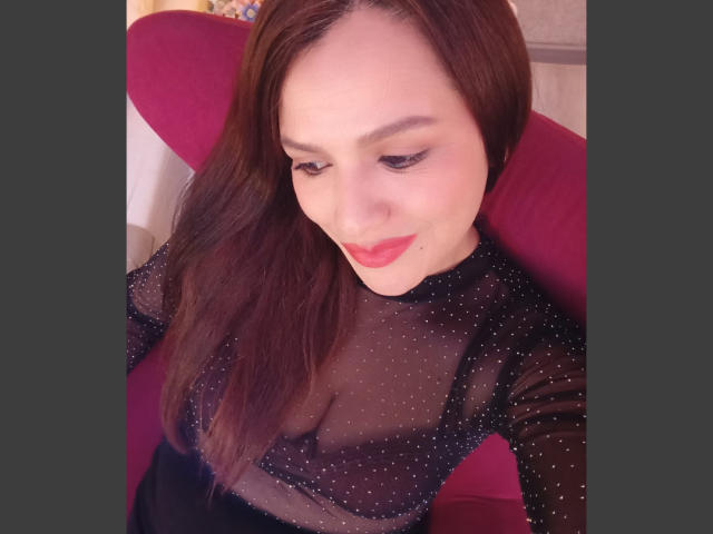 CameronViviane, avec du rouge à lèvres, portant un haut noir transparent avec de petits points blancs, dévoilant ses seins, assise sur une chaise rouge, regardant vers le bas, avec des cheveux bruns..