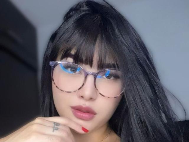 LunaAndKlaus, avec ses cheveux noirs, ses lunettes, son rouge à lèvres rouge et son anneau de nez, touchant son menton avec ses ongles rouges, montrant un tatouage sur son doigt..
