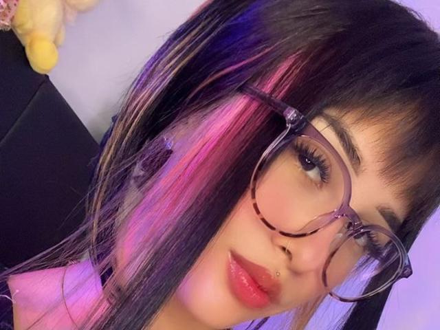 LunaAndKlaus has dark hair with purple and pink highlights, wears large tortoiseshell glasses, and has a nose piercing with glossy pink lips. She's a cute, sexy chick.