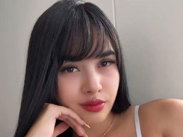 LunaAndKlaus has long black hair, brown eyes, and red lipstick, with a nose ring and a gold necklace, resting her chin on her hand, showing off her painted nails.