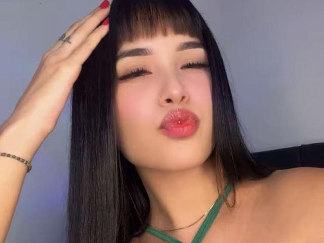 LunaAndKlaus tem lábios rosados e carnudos, olhos fechados e cabelo preto liso com franja. Ela está chupando o lábio superior, tocando a cabeça com os dedos vermelhos e usando um top verde de alças..