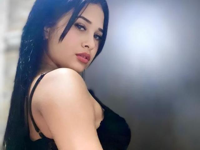 LunaAndKlaus mit langem, schwarzem Haar, rotem Lippenstift und einem schwarzen BH, die ihre nackte Schulter und ihr leicht sichtbares Dekolleté zeigt, blickt über ihre Schulter mit einem sexy, selbstbewussten Ausdruck..