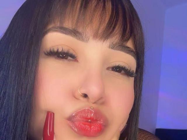 LunaAndKlaus a les cheveux noirs et lisses, un anneau de nez et des lèvres rouges brillantes avec un piercing. Elle tient un vibreur rouge près de sa bouche. Elle a de longs cils épais..