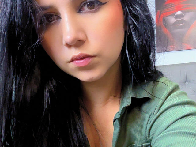 Kkatalina's face with dark, wavy hair, green shirt, and winged eyeliner. She has a serious expression, pink lips, and a cute nose. Her tits are partially visible.