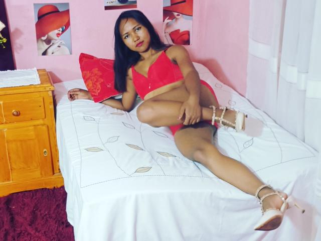 KiasimVioletta, Latina, with long black hair, wearing red lace bra and panty set, white high heels, on bed, leg raised, showing her pussy, red pillow, pink walls.