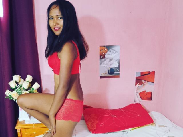 KiasimVioletta with dark skin, black hair, small red lace bra, and matching red lace panties, kneeling on bed, showing her ass, smiling, with white roses to the left.
