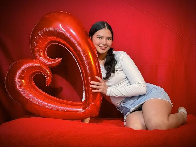 LizzieSweets, with black hair in pigtails, holds a shiny red heart balloon, kneeling on a red surface, wearing a white long-sleeve top and denim shorts.
