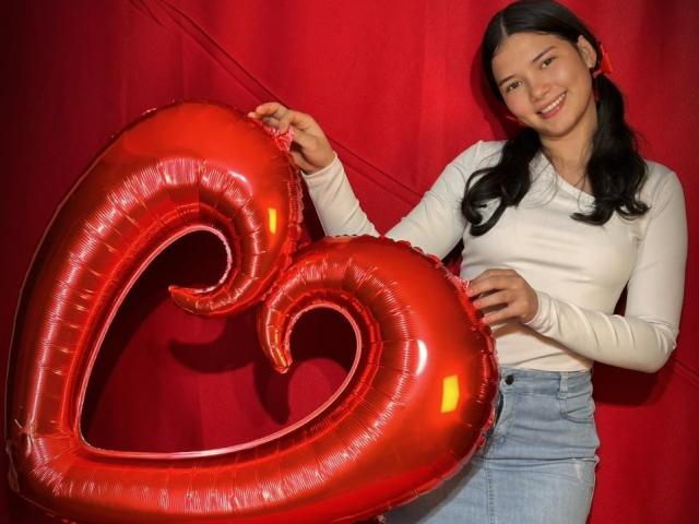 LizzieSweets tient des ballons en forme de cœur rouges brillants, elle porte une chemise blanche à manches longues et un jean bleu, et elle sourit avec ses couettes foncées. Ses fesses ne sont pas visibles..