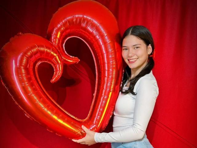 LizzieSweets tenant un ballon rouge en forme de cœur, souriant avec des couettes noires, portant une chemise blanche à manches longues et un jean bleu, sur un fond rouge..