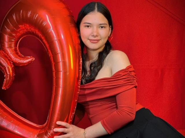 Lizzie, vêtue d'un haut rouge à une épaule, d'un pantalon noir, tenant un ballon rouge brillant, souriante, avec de longs cheveux noirs, sur un fond rouge, décolleté visible, mignonne, sexy, éclairage doux, pose détendue..