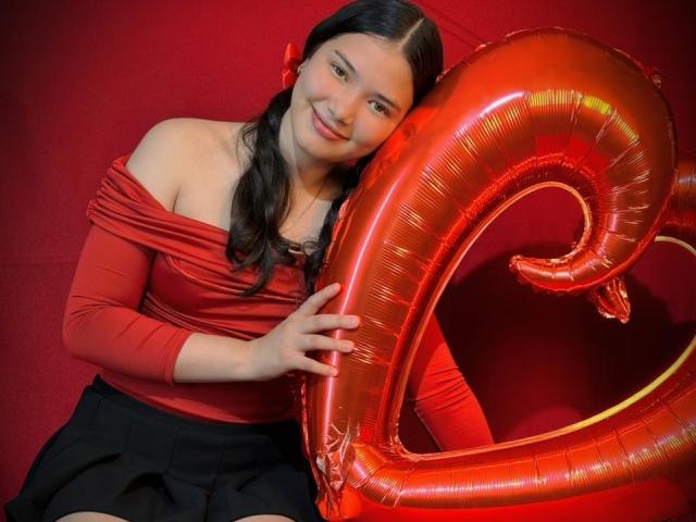 LizzieSweets with long black hair in pigtails, red off-shoulder top, and black skirt, holding shiny red "10" balloon, smiling at camera. Boobs slightly visible under top.