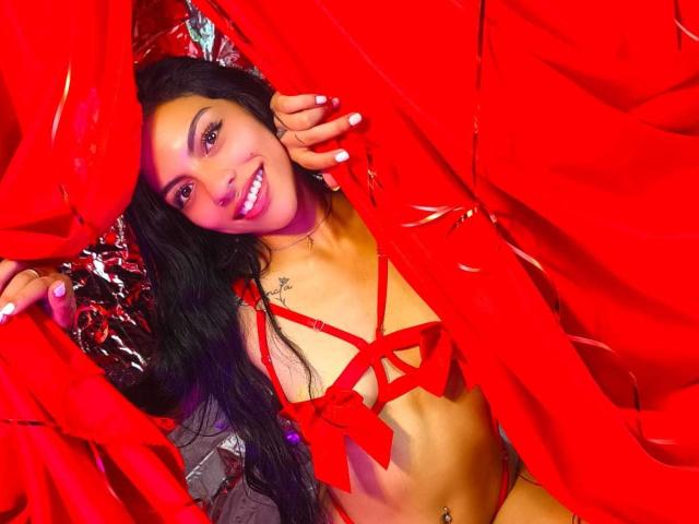 RoxannaMoretti with long black hair, smiling, wearing red strappy lingerie, holding red fabric, exposing small tits, tattoo on right shoulder, and a belly button piercing.
