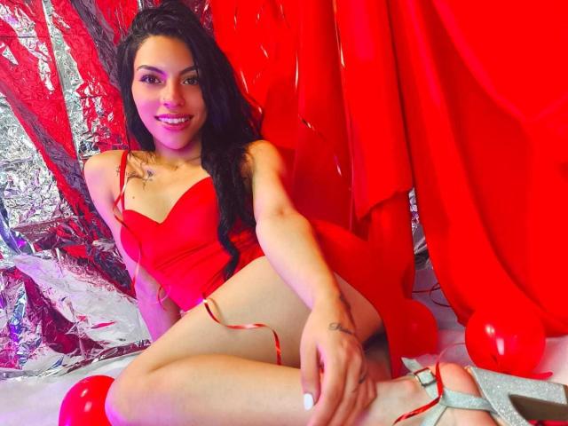 RoxannaMoretti with long black hair, red dress, and red heels, smiling, legs crossed, showing her ass, red background, and silver foil, tattoos on her right arm and left thigh.