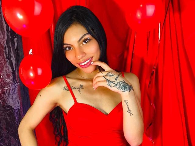 RoxannaMoretti with long black hair, red dress, and tattoos on chest and hand, smiling, finger to lips, red balloons and red background, showing her tits and cute ass.