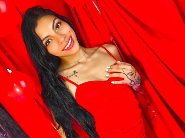 RoxannaMoretti with long black hair, red dress, white nails, tattoos on chest and hand, smiling, holding dress strap, red background. Boobs visible, sexy, cute, attractive.