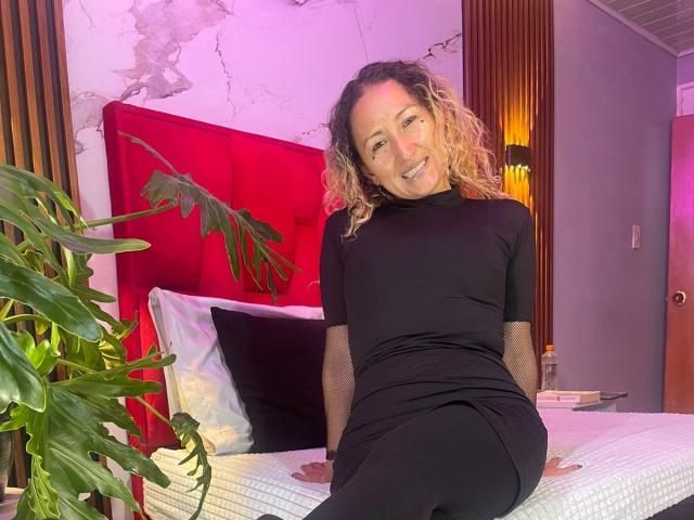 SweeettCharlotte with curly blonde hair, black dress, and mesh sleeves, sitting on a bed with a red headboard, smiling, green plant on the left, purple lighting.