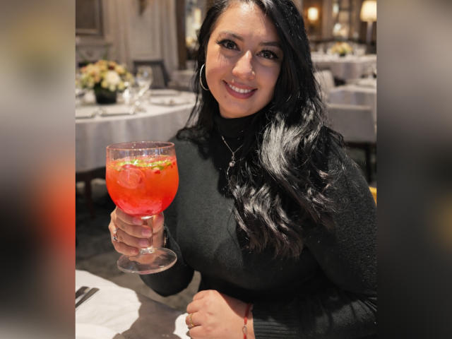 AshleeyPrinces with long black hair, wearing a black turtleneck, holding a red cocktail with green garnish, smiling at the camera, in a dimly lit restaurant.