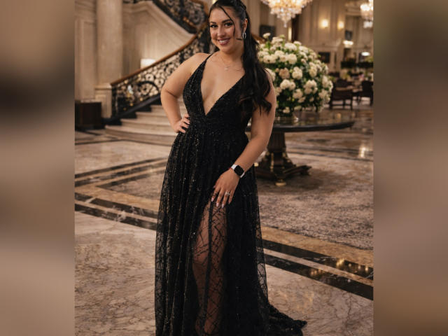 AshleeyPrinces in a black, deep V, sequined gown with a high slit, standing in an elegant room, wearing a black bracelet, smiling, long dark hair, large tits.