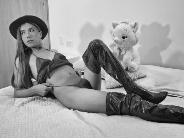 MissMelissaHot, avec son chapeau noir, son haut transparent et ses bottes brillantes montantes, est allongée sur le lit avec un jouet en forme de cochon, exposant ses petits seins plats et son corps mince et sexy..