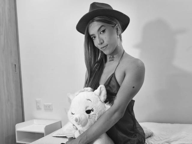MissMelissaHot com cabelo comprido, usando um chapéu e um vestido, segurando um porquinho de pelúcia com a mão em seu traseiro, em pé ao lado de uma cama em uma foto em preto e branco..