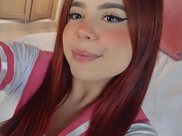 MelaniWare has straight, medium-length red hair, a light skin tone, and subtle makeup with black eyeliner. She's wearing a pink and silver top, and her lips are slightly parted in a small smile. Her eyebrows are dark and well-defined. The image has a soft, warm light. The focus is on her face, which is centered in the frame. The texture of her hair is smooth and shiny. Her eyes are slightly upturned, giving a gentle, approachable look. The overall composition is simple and close-up.