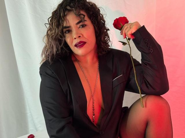 AbriilBeltran with curly brown hair, wearing a black blazer, deep cleavage, holding a red rose, sitting, red lipstick, necklace, white and red background, slightly smug expression.