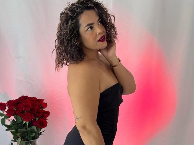 AbriilBeltran with curly brown hair, dark red lipstick, black strapless dress, right hand on neck, red roses on left, pink and white background, subtle tattoo on right arm.