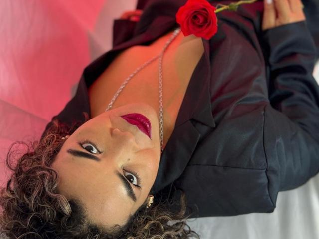 AbriilBeltran with curly brown hair, wearing a black shirt, red lipstick, and a necklace, lying down with a red rose on her chest, looking up.