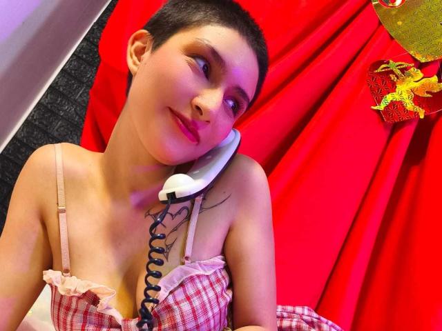 IsabellaHallad with short dark hair, pink checkered top, small tits, holding white phone, red curtain, black tattoo on right shoulder, slight smile, pink lipstick, dark eyes.