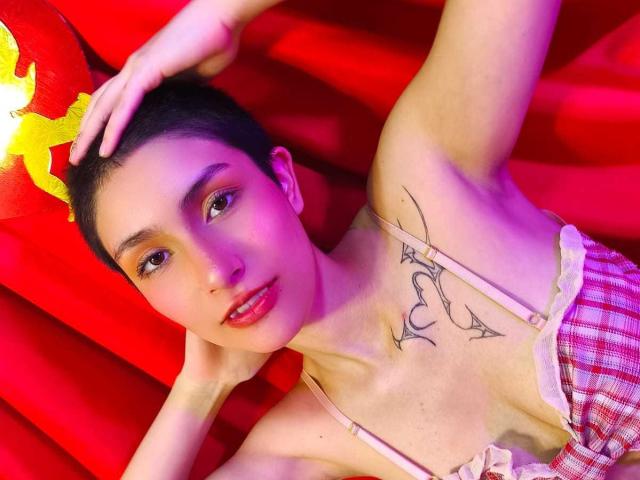IsabellaHallad with short black hair, red lipstick, and a tattoo under her tits, lying on red fabric, wearing a pink and white checkered top, hand on head.