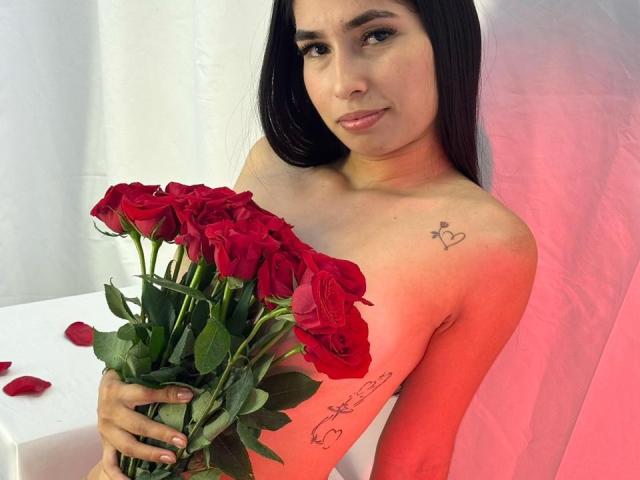 NatashaFerrec, avec ses cheveux foncés, tenant un bouquet de roses rouges, cachant ses seins avec son bras. Elle a un petit tatouage en forme de cœur sur son épaule droite..