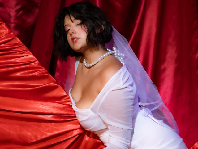 MisakiWong with short black hair, pearl necklace, white sheer dress, and veil, leaning on red satin, showing her tits. Red background, glossy texture. Soft, sensual, confident pose.