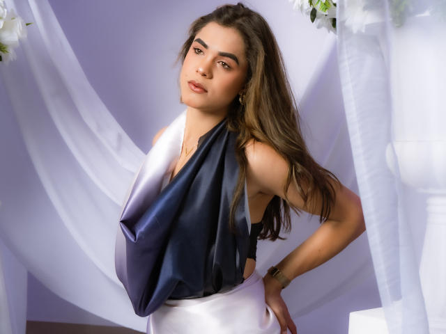 AliceCoolenk with long brown hair, wearing a shiny blue and white dress, showing her side boob and looking at the camera, standing with her hand on her hip.