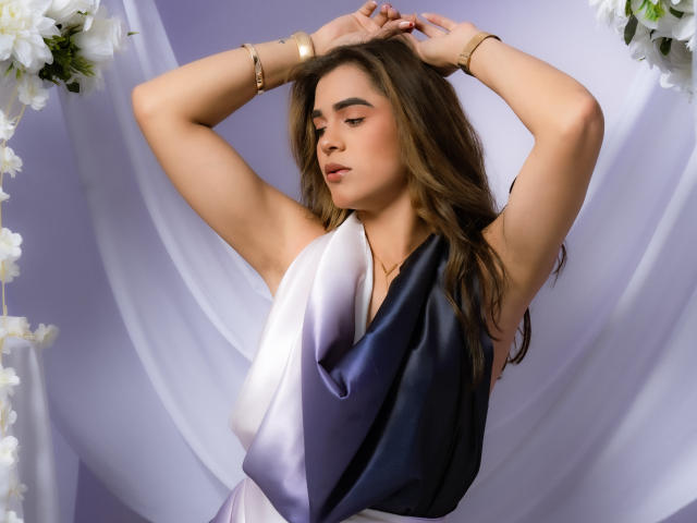 AliceCoolenk with arms raised, wearing a gradient dress from black to white, brown hair, soft makeup, gold bracelets, looking down, white flowers on left. She's got nice tits.