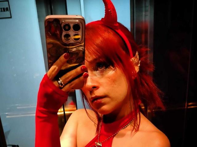 LolyYunno has red hair, wears a red devil horn headband, and a matching choker. She has red nail polish and devil ears, with a serious expression. She's holding a phone with a reflective case, taking a mirror selfie. Her right hand has a red glove, and she's wearing a silver ring. The lighting is warm, highlighting her fair skin and freckles. The mirror shows reflections of the room. The overall style is casual and slightly provocative.