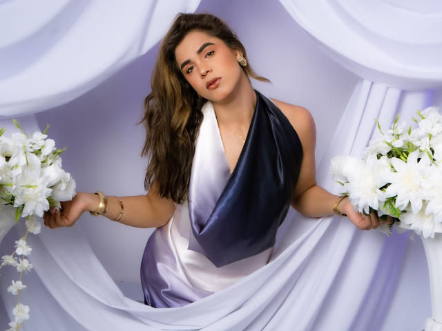 AliceCoolenk stands in the center, wearing a sleek, gradient dress that transitions from dark blue to white. She holds white flower bouquets in both hands. Her long, wavy brown hair cascades over her shoulder. She has a neutral expression, with subtle makeup emphasizing her dark eyes and full lips. Gold earrings and a bracelet add elegance. The white, draped fabric in the background contrasts with her dress, enhancing the visual impact. The image uses soft lighting and a slight tilt, emphasizing AliceCoolenk's relaxed yet confident posture.
