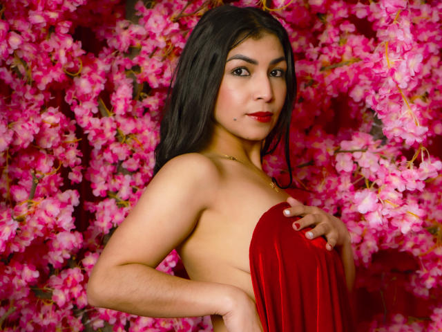 ChanelBrouw with dark long hair, red lipstick, and medium tits, covering her bare back with a red velvet cloth, standing against a pink floral background.