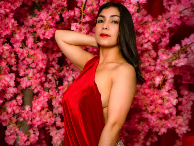 ChanelBrouw with dark hair, red velvet dress, sideboob, arm raised, looking at camera, pink cherry blossom background, red lipstick, sexy, confident.