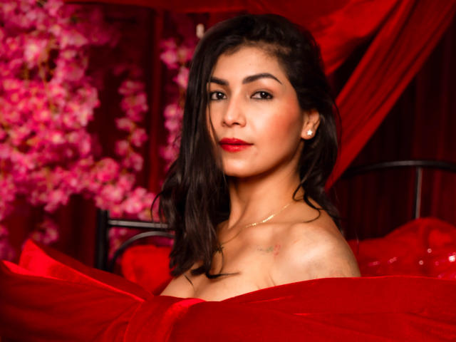 ChanelBrouw, a dark-haired woman with medium-length wavy hair, stands in a red setting. She has a medium skin tone, arched eyebrows, and wears bright red lipstick. Her eyes are slightly downcast, giving a subtle, confident look. She's dressed in a red, off-shoulder, silky dress that partially reveals her shoulders. She has small pearl earrings and a thin gold necklace. The background is blurred with pink floral patterns, contrasting with the red. The overall composition is bold and sensual, with rich red colors dominating the image.