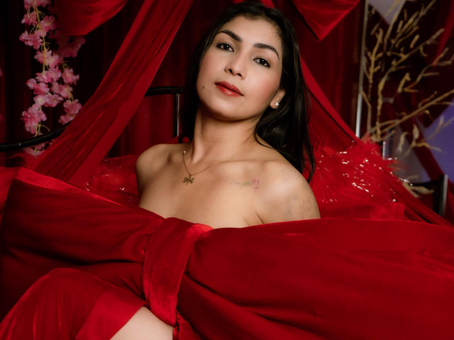 ChanelBrouw with medium-length black hair sits in a red, velvety, and sequined setup. She has light olive skin, small earrings, and a gold necklace with a star pendant. Her red lipstick matches the rich red fabric that surrounds her. Her bare shoulders show a small tattoo on her right collarbone. Pink cherry blossoms and gold branches are partially visible in the background. The composition emphasizes red and velvet textures. The lighting is soft, highlighting her facial features and the shiny fabric. The image has a watermark in the bottom left corner.
