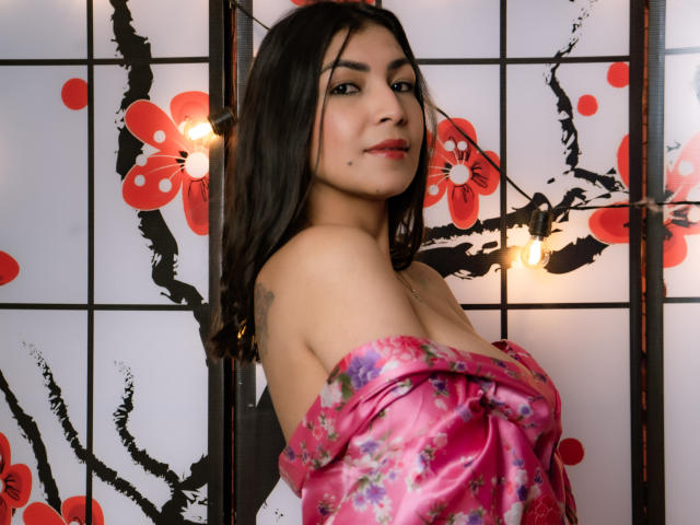 ChanelBrouw with dark hair, red lipstick, and a pink floral kimono, exposing her bare shoulders and small tits, standing in front of a Japanese-style screen with red flowers.
