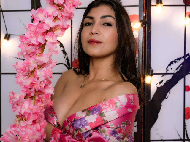 ChanelBrouw with pink floral kimono, holding pink cherry blossom branch, showing big tits, dark brown hair, red lipstick, green necklace, cute smile, soft light, warm colors.